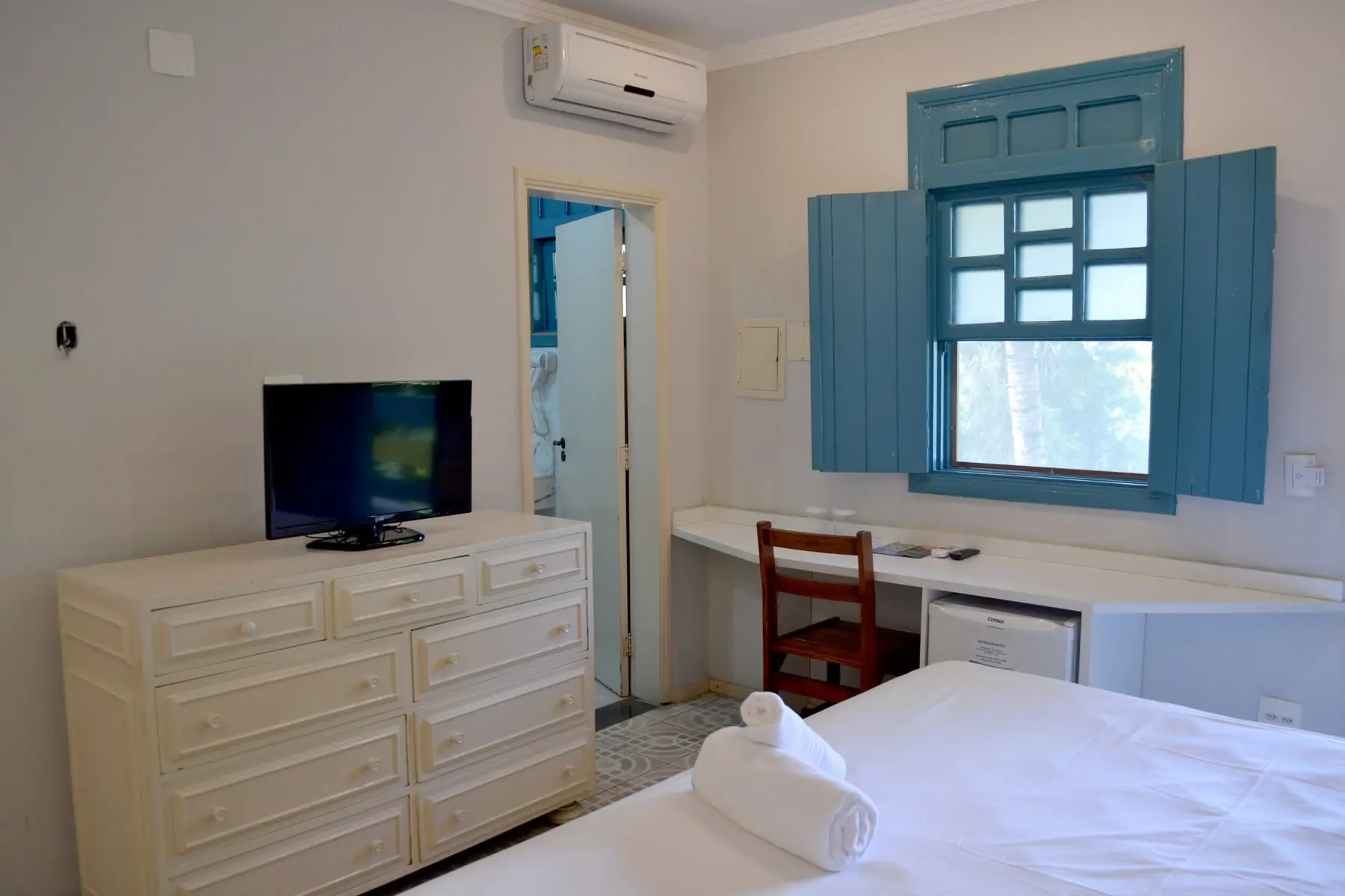 Quarto de acomodação colonial do Spa Med, com janela de madeira azul, cômoda clara e televisão