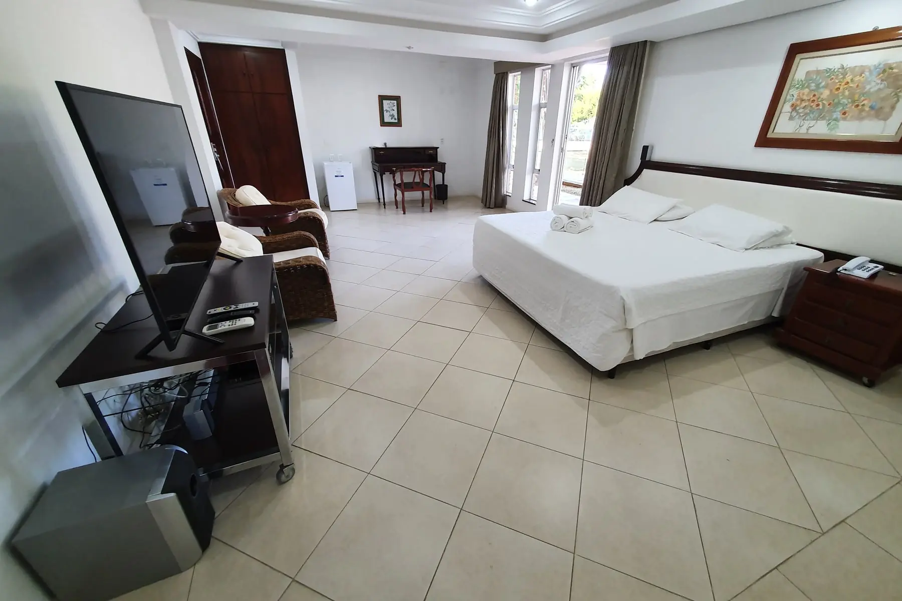 Quarto amplo com cama de casal, poltronas de vime, televisão e porta de vidro para a varanda