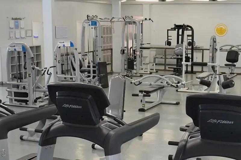 Academia com fileira de esteiras e aparelhos de musculação, iluminada por luminárias corridas