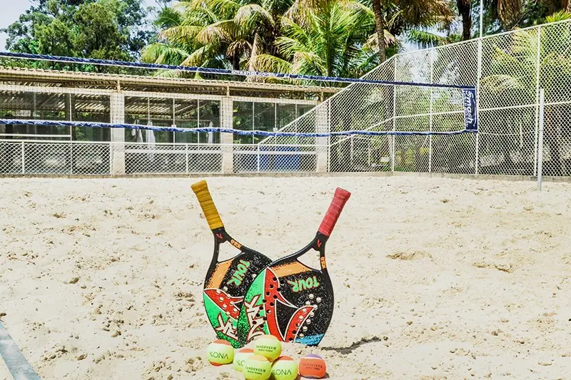 Raquetes de beach tennis apoiadas na areia, com a quadra e a rede ao fundo