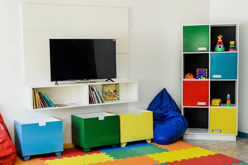 Brinquedoteca com tapete emborrachado colorido, estante de brinquedos, pufe e televisão