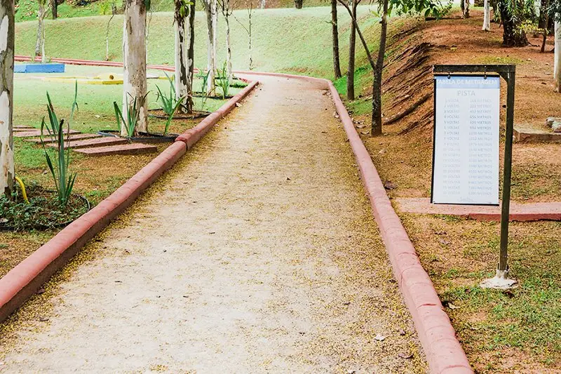 Pista de caminhada de saibro contornando o gramado, com placa de sinalização e mata em volta