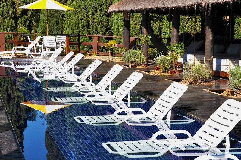 Fileira de espreguiçadeiras brancas à beira da piscina, sob a palhoça e um guarda-sol amarelo