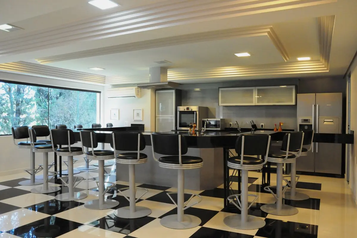 Espaço gourmet com bancada e banquetas pretas, piso xadrez e janela para o verde