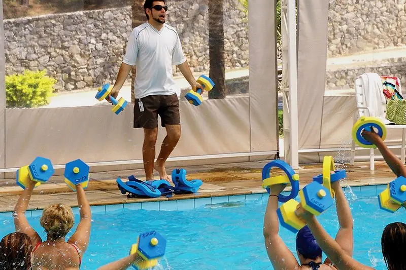 Turma de hidroginástica na piscina com halteres de espuma, e o professor orientando da borda