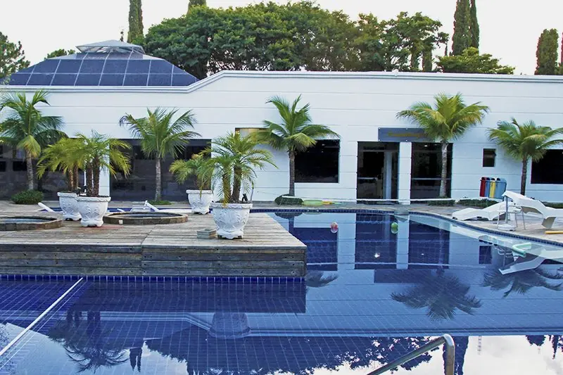 Piscina de hidroterapia coberta, vista do deck, com o bloco branco e as palmeiras ao fundo