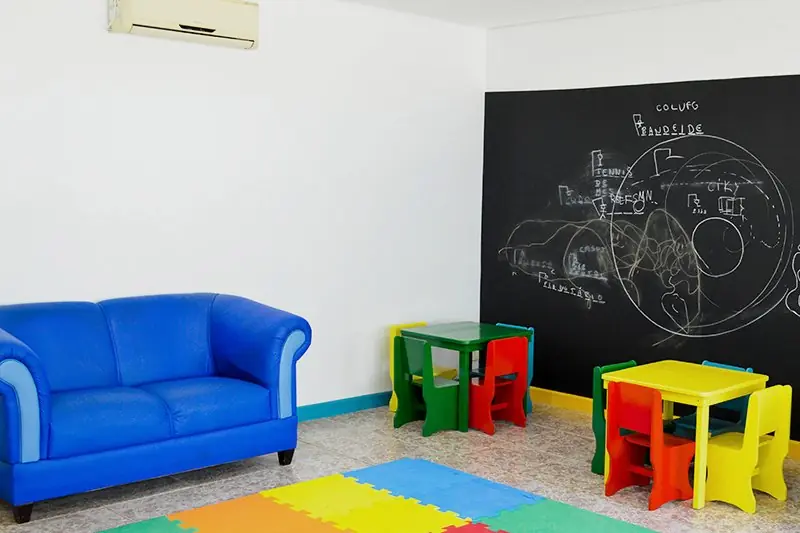 Espaço kids com sofá azul, mesinhas coloridas, tapete emborrachado e lousa na parede