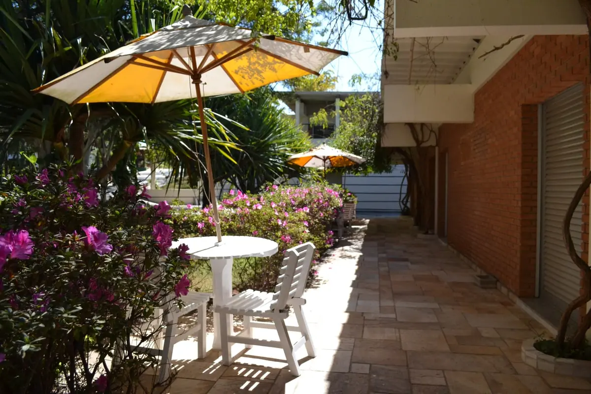 Pátio com mesa branca e guarda-sol amarelo entre os blocos, com flores em primeiro plano