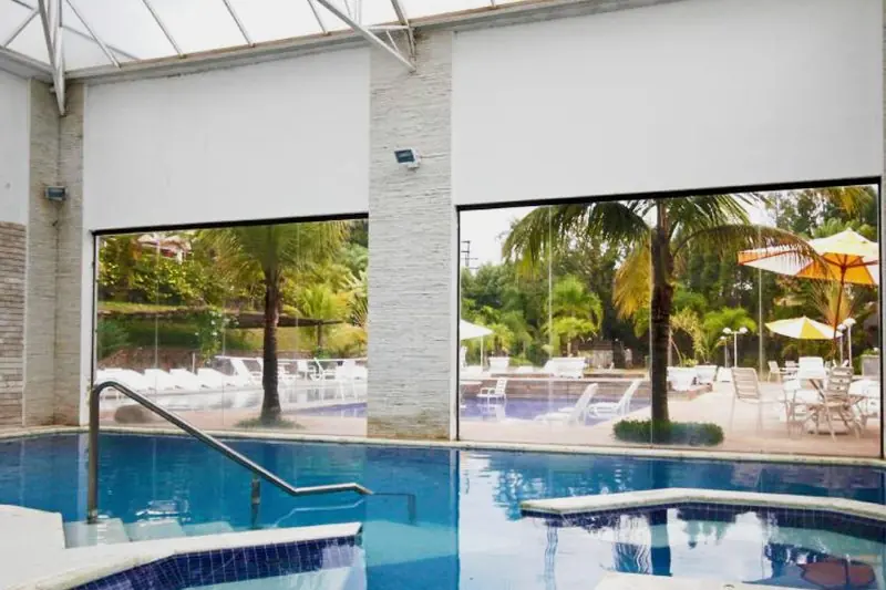 Piscina coberta sob estrutura de vidro, com a piscina externa visível pela parede envidraçada