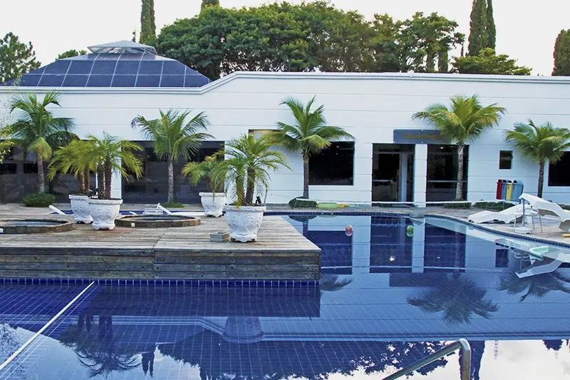 Piscina externa em L com água azul, palmeiras em volta e o bloco da sede ao fundo