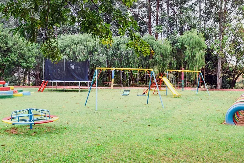 Playground no gramado, com balanços, escorregador, gangorra e gira-gira, cercado de árvores