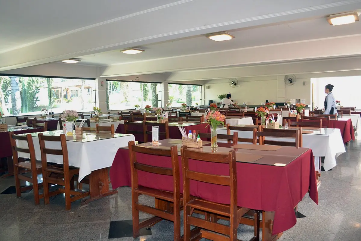 Salão do restaurante com mesas postas de toalha branca e sobretoalha vermelha, e equipe atendendo