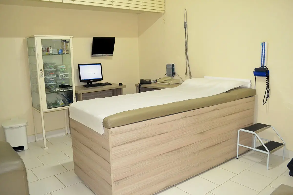 Sala de exames com maca, armário de vidro, monitor e equipamento de parede