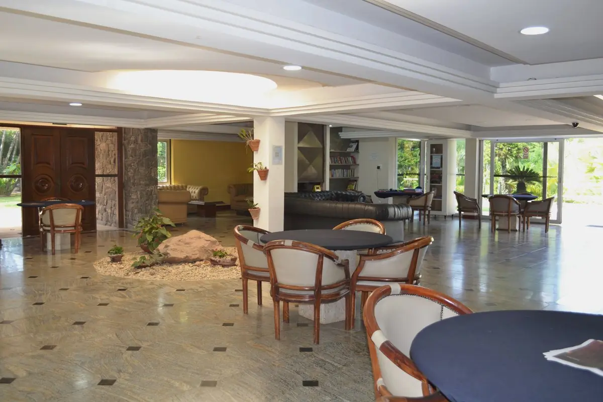 Salão social com mesas redondas, poltronas e porta de vidro para o jardim