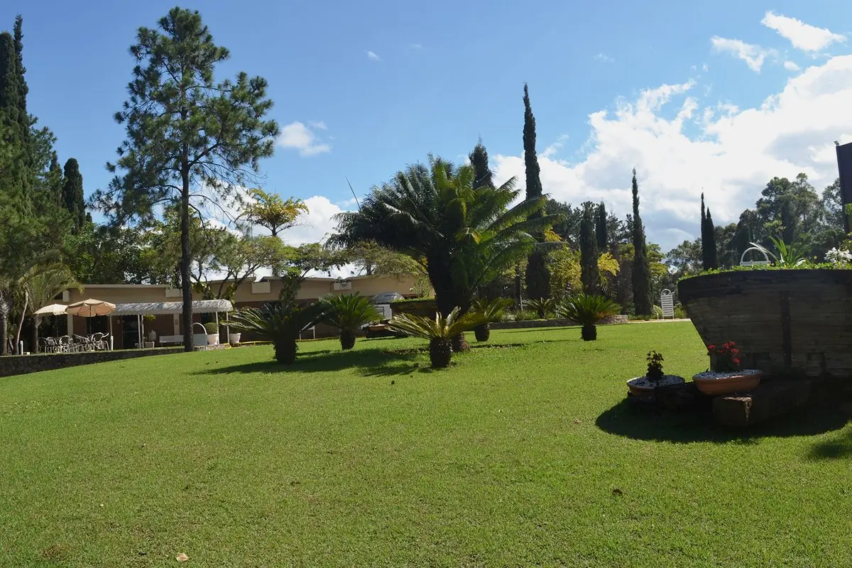 Gramado aberto em frente à casa sede, com árvores altas e um jardim de pedra em primeiro plano