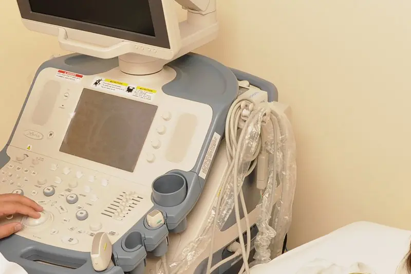 Profissional operando o aparelho de ultrassom ao lado da maca, com o monitor à frente