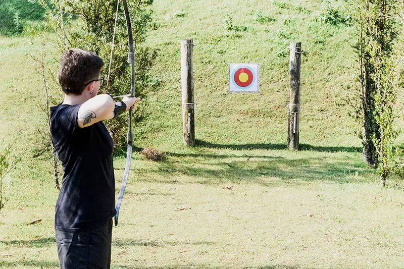 Praticante de arco e flecha de perfil, com o alvo à frente e o gramado subindo ao fundo