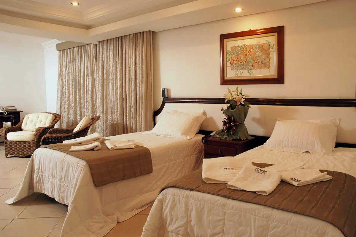Interior da Suíte Master com duas camas, poltronas e cortina fechando a janela